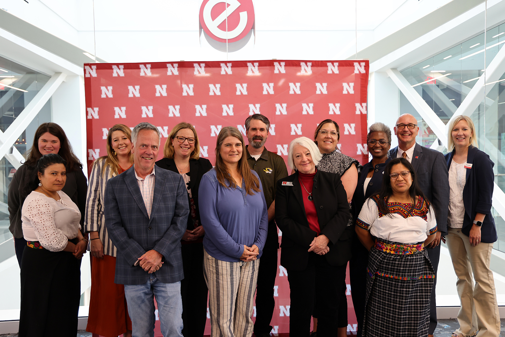 Thirteen people pose for a photograph in two rows, in front of a Husker "N" backdrop.