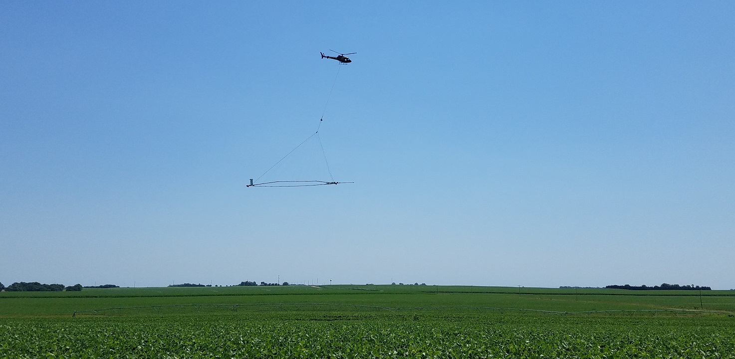 A helicopter carries a large loop of wire whose electromagnetic signaling enables extensive imagery of subsurface conditions and the creation of a 3D geological model.