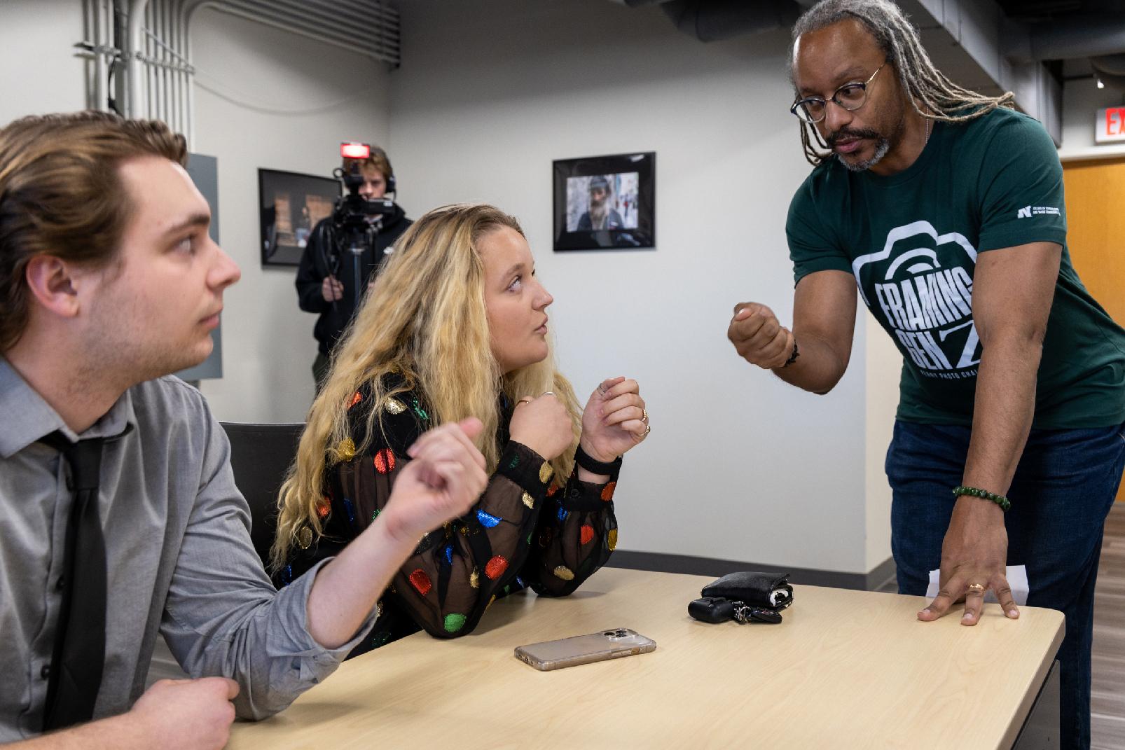 Shoun Hill, standing, talks to students Hayden Rooney and Merideth Gamet, seated at a table, while a cameraman films in the background.