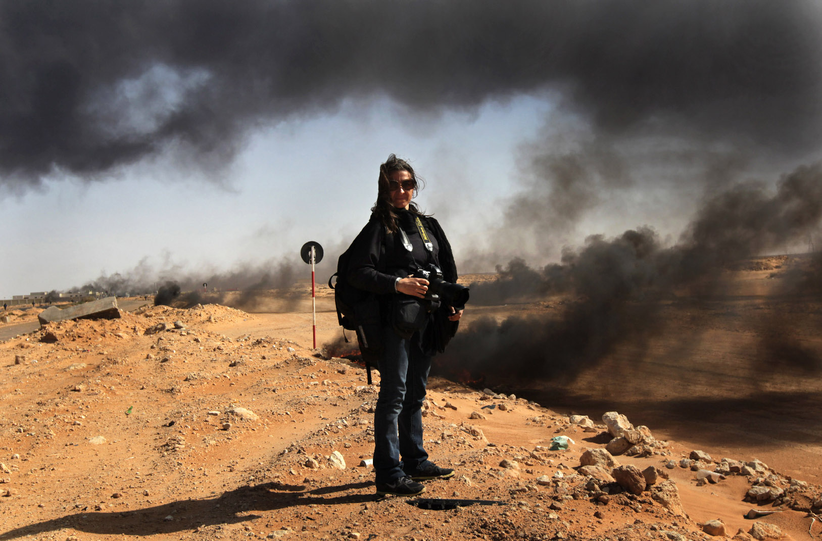 Lynsey Addario stands with a camera on rocky terrain, with plumes of black smoke in the background.