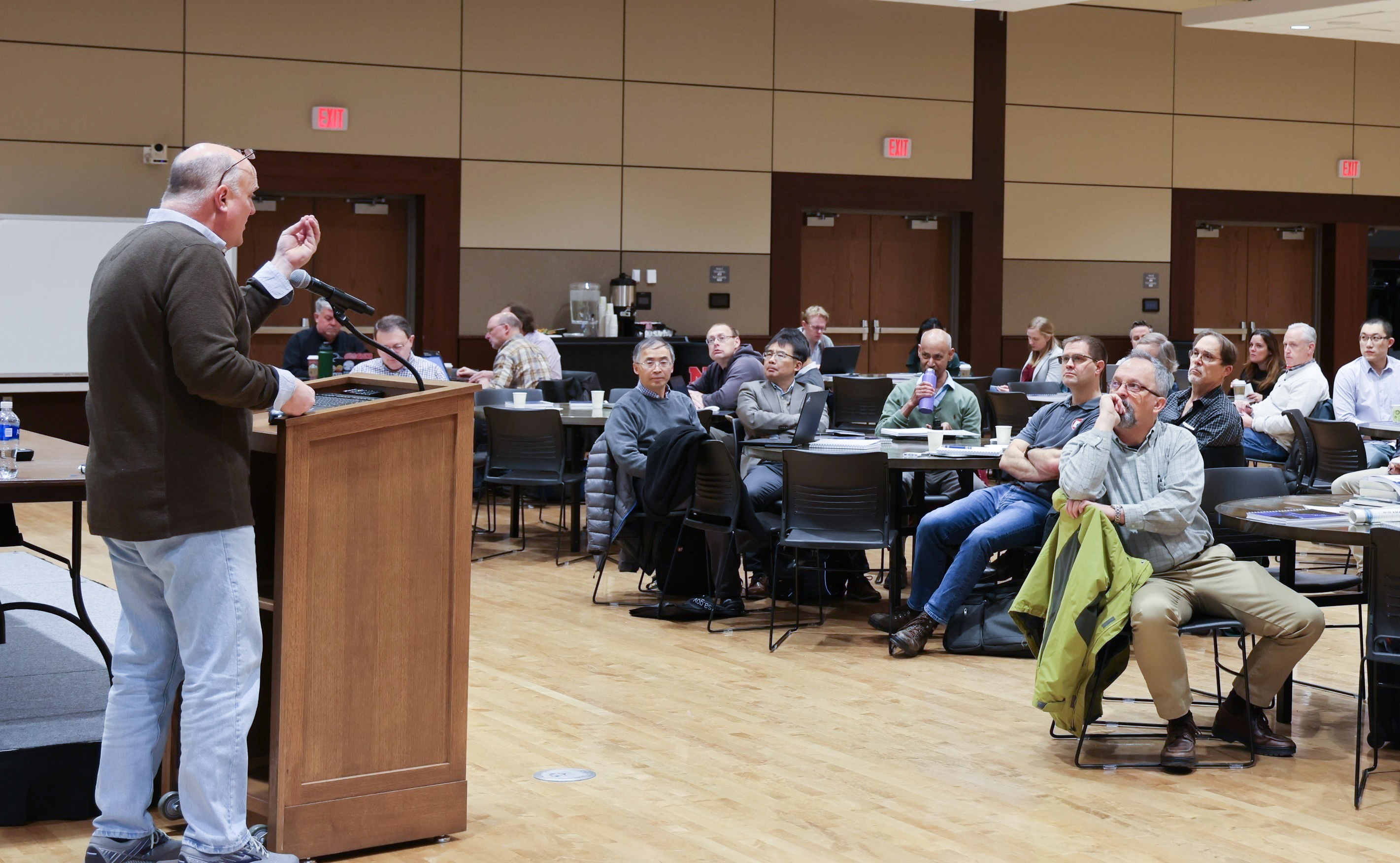 A man at a lectern speaks to about 20 people seated around tables.