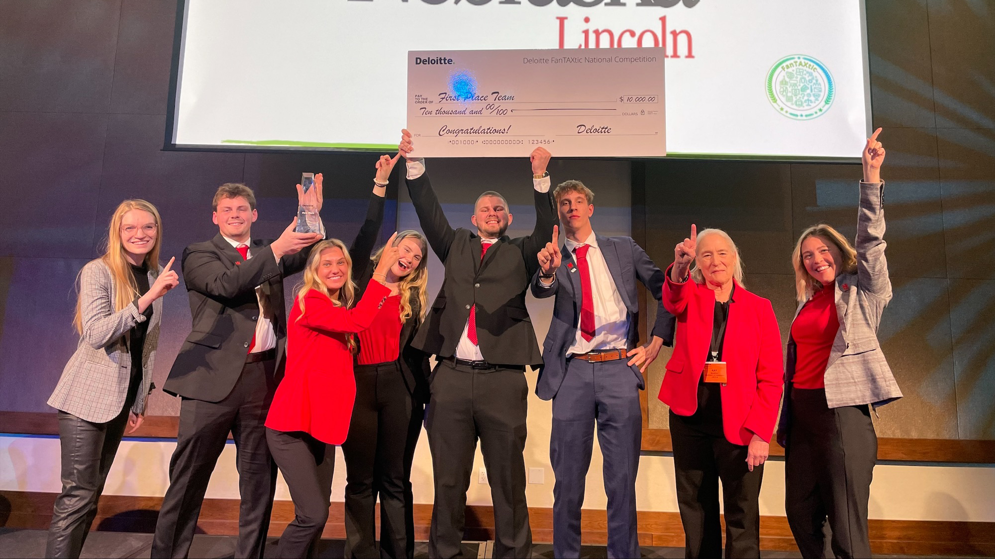 Seven students and their adviser pose with a giant check and a glass trophy, holding up their index fingers to signal that they are "No. 1."