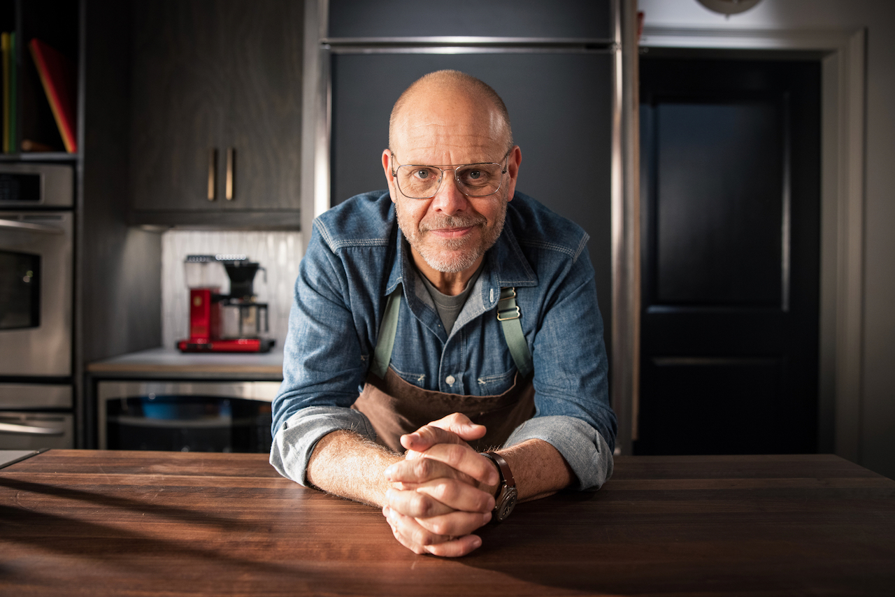 Alton Brown, wearing a chef's apron, leans over a counter, with his hands crossed in front of him.