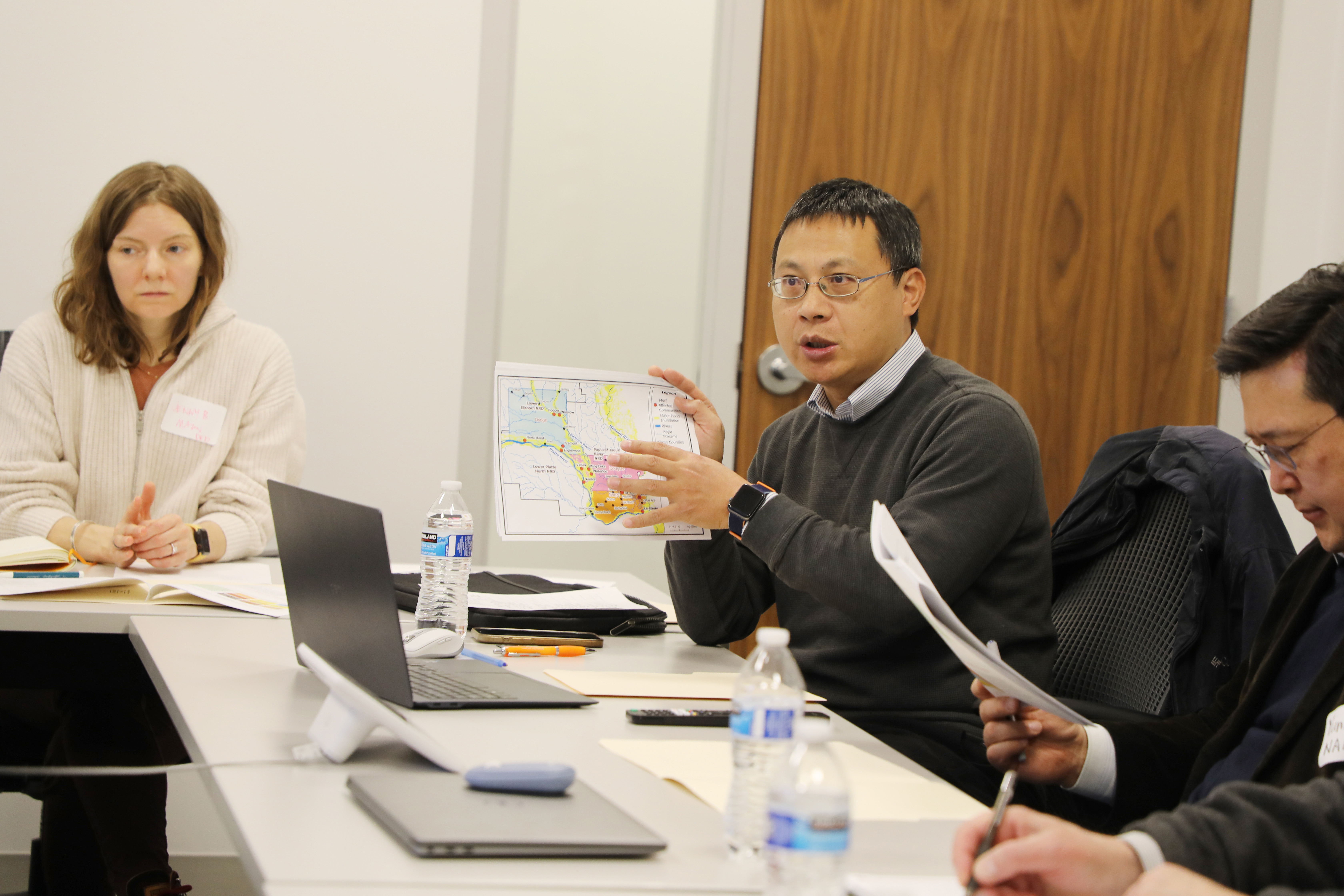 Jenny Mason, Zhenghong Tang and Yunwoo Nam sit at meeting tables. Tang holds a map.