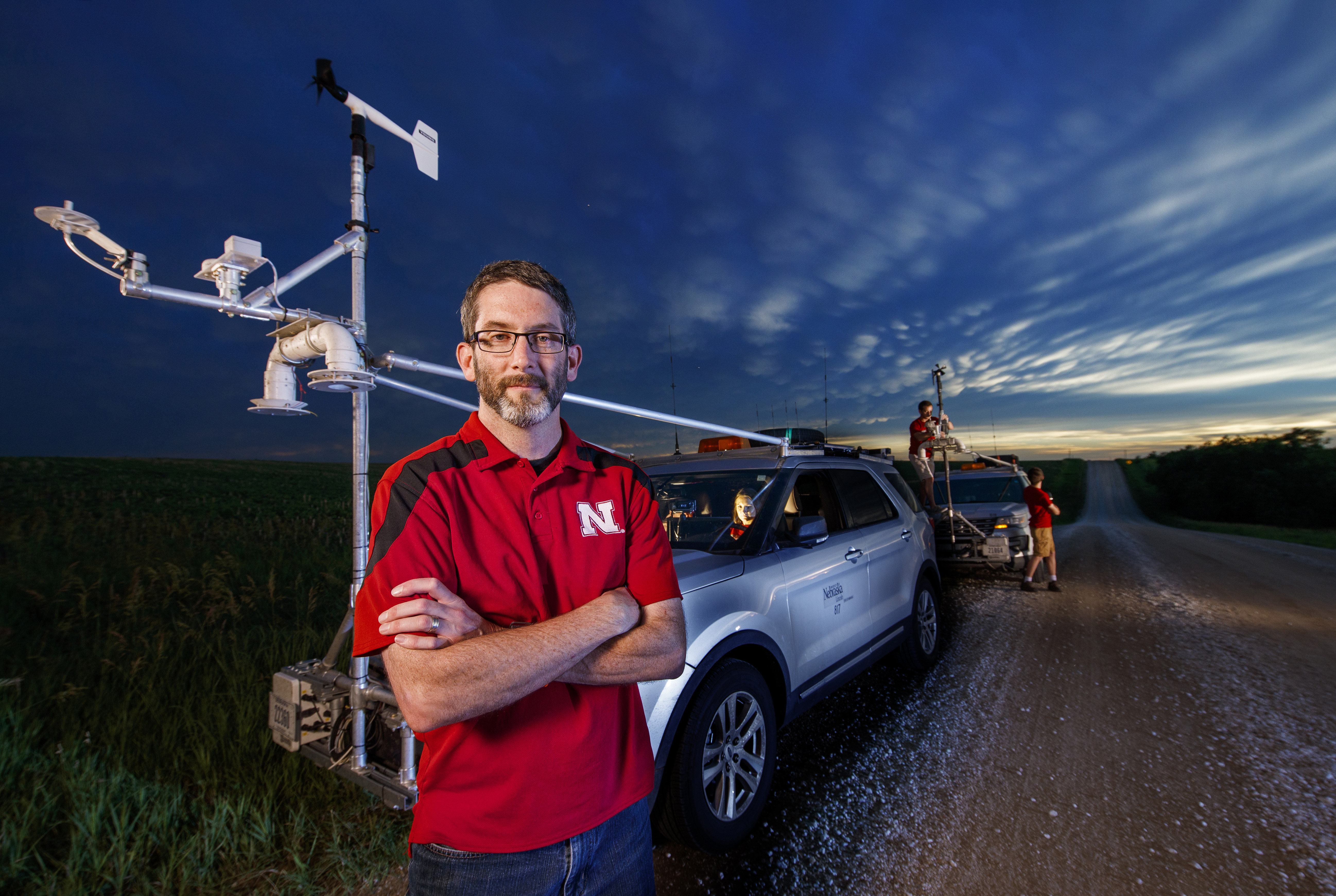 Adam Houston stands with his arms crossed in front of two storm-chasing vehicles.