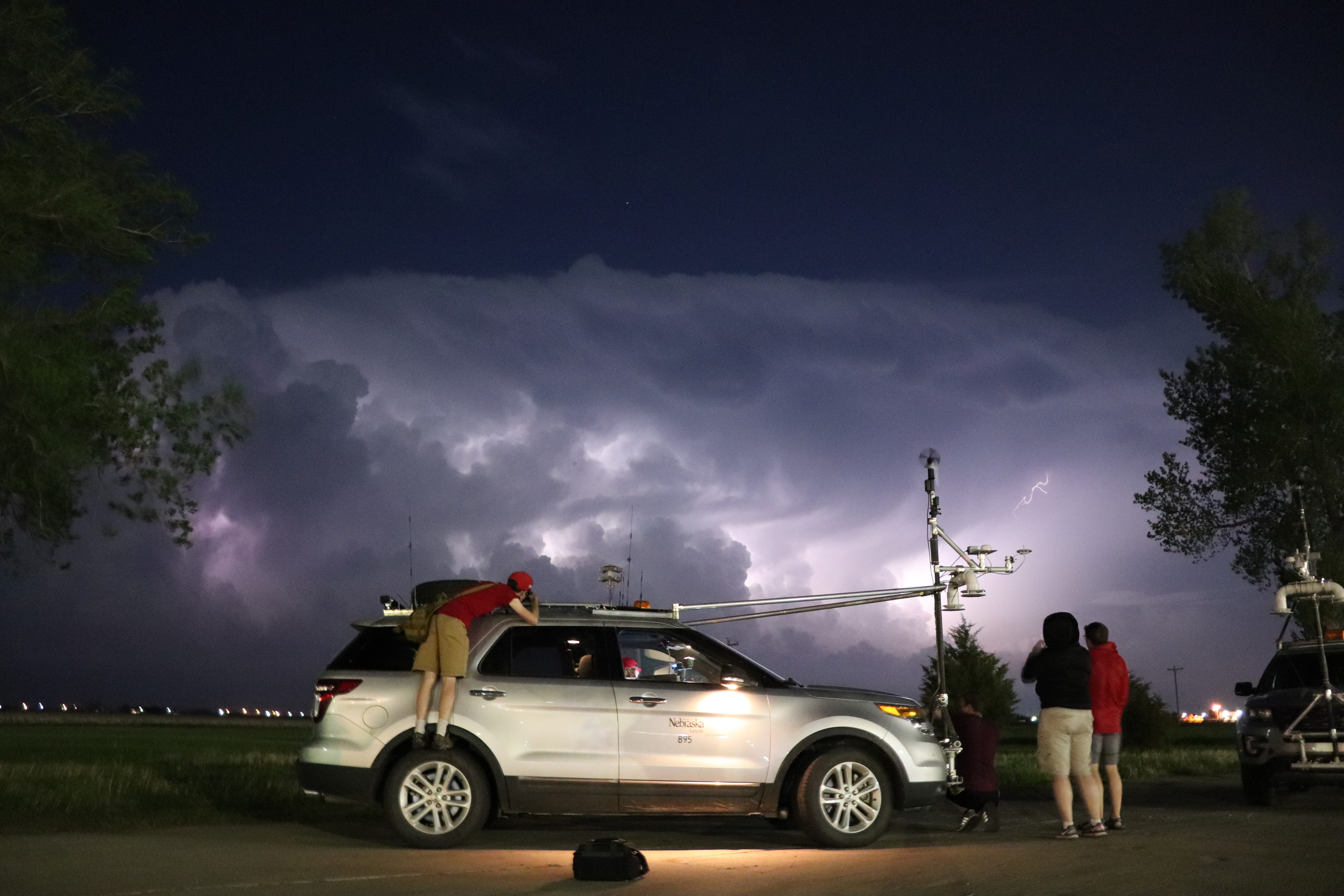 Four people stand around a storm-chasing vehicle at night, with storm clouds in the background.