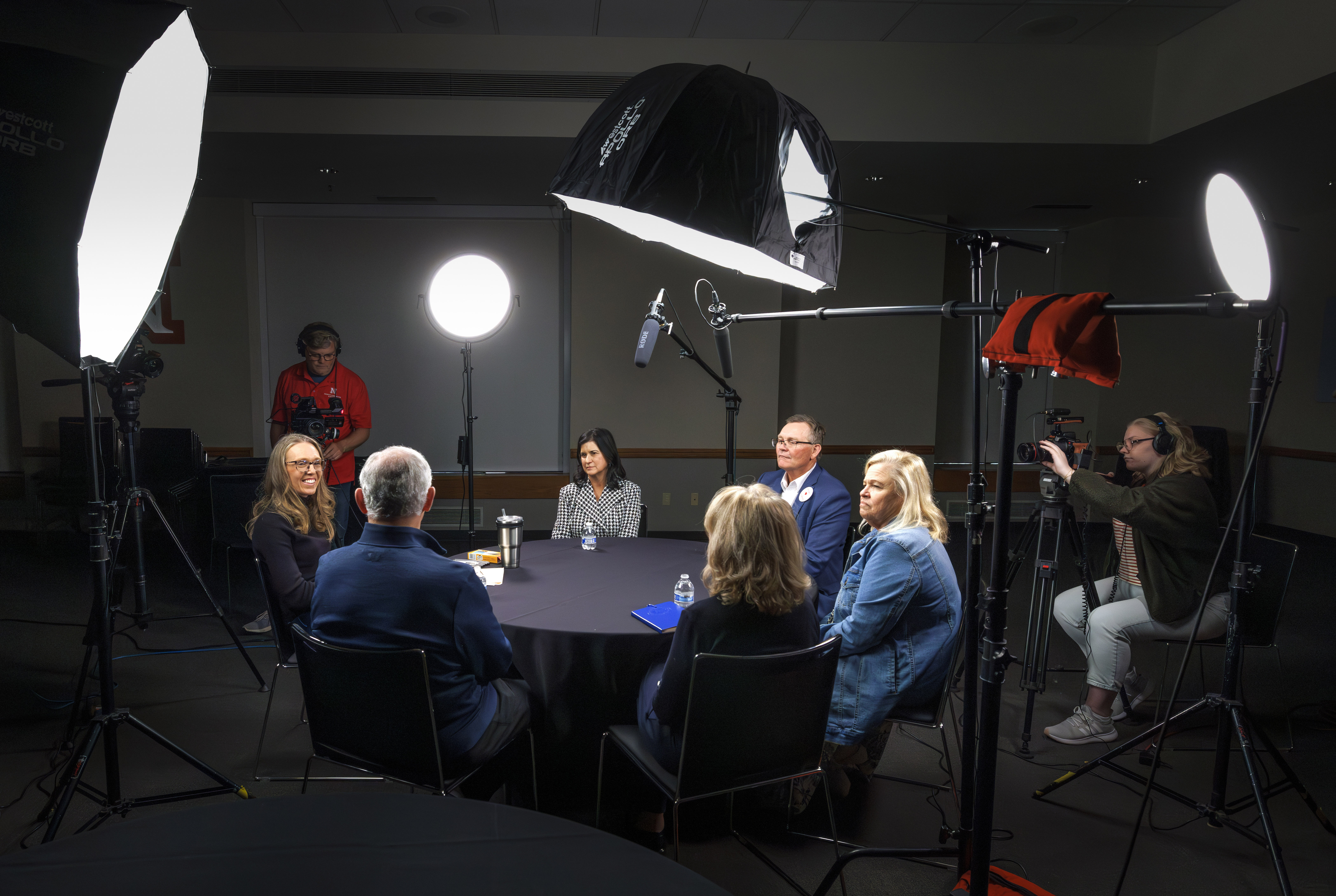 Four women and two men sit at a round table in a dark room, surrounded by lighting umbrellas, as a man and woman film.