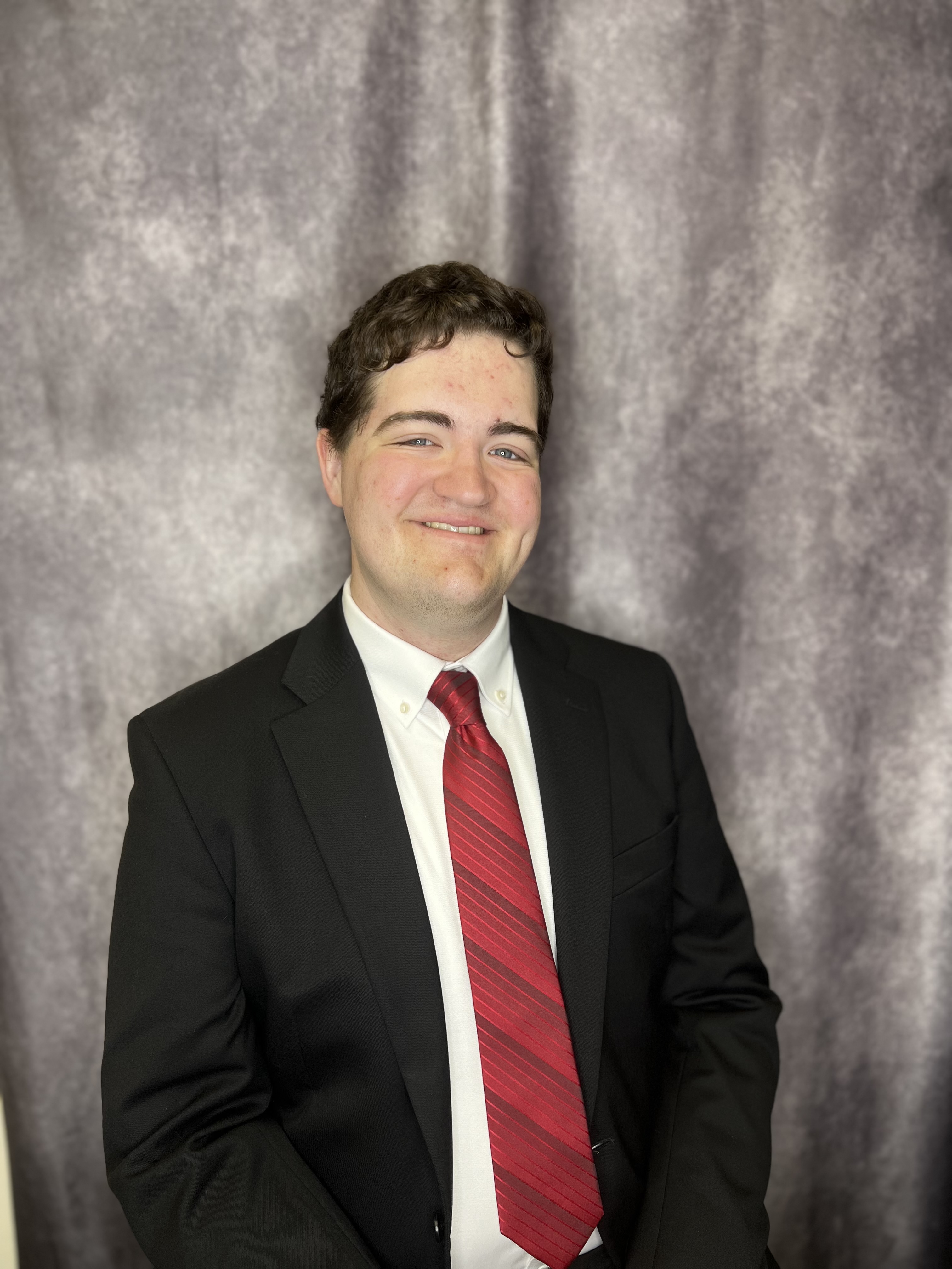 Color portrait of Carson Haberyan, wearing a black suitcoat and white dress shirt with red tie, against a gray background.