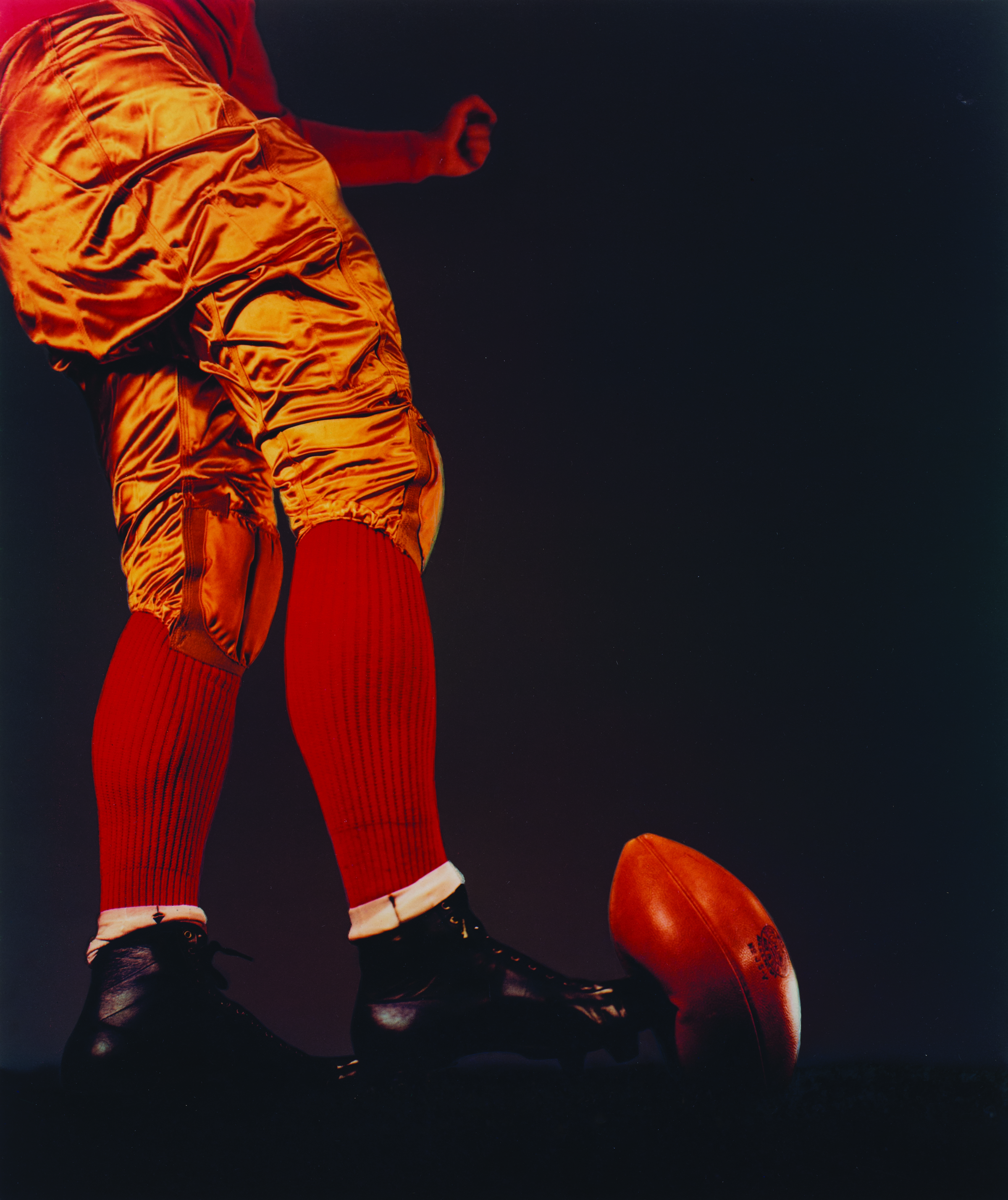 A football player wearing gold pants, red socks and black cleats kicks a football against a black background in Harold Edgerton's "Football Kick."