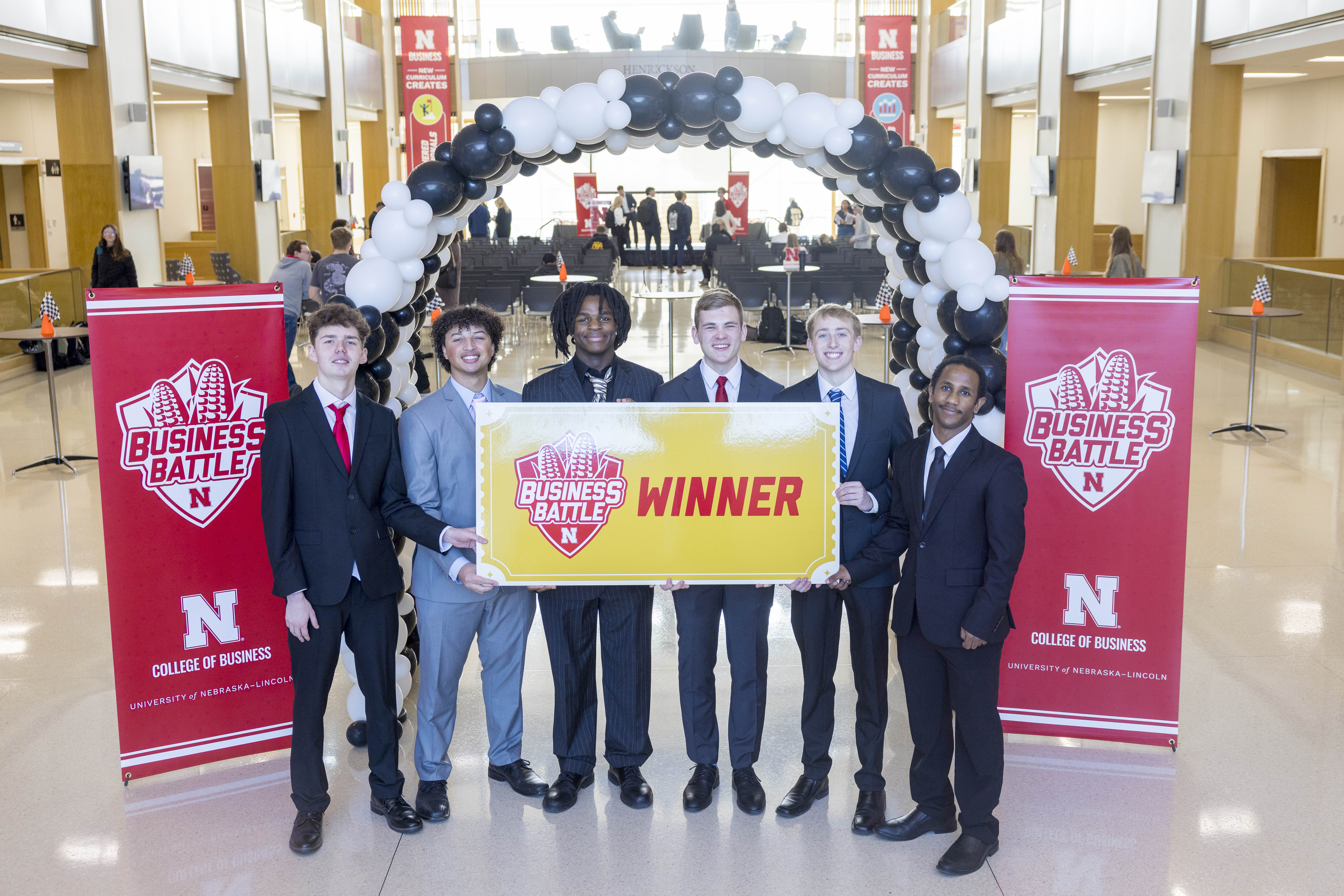 Six young men in suits pose with a poster that reads "Business Battle Winner" in the Howard L. Hawks Hall atrium.