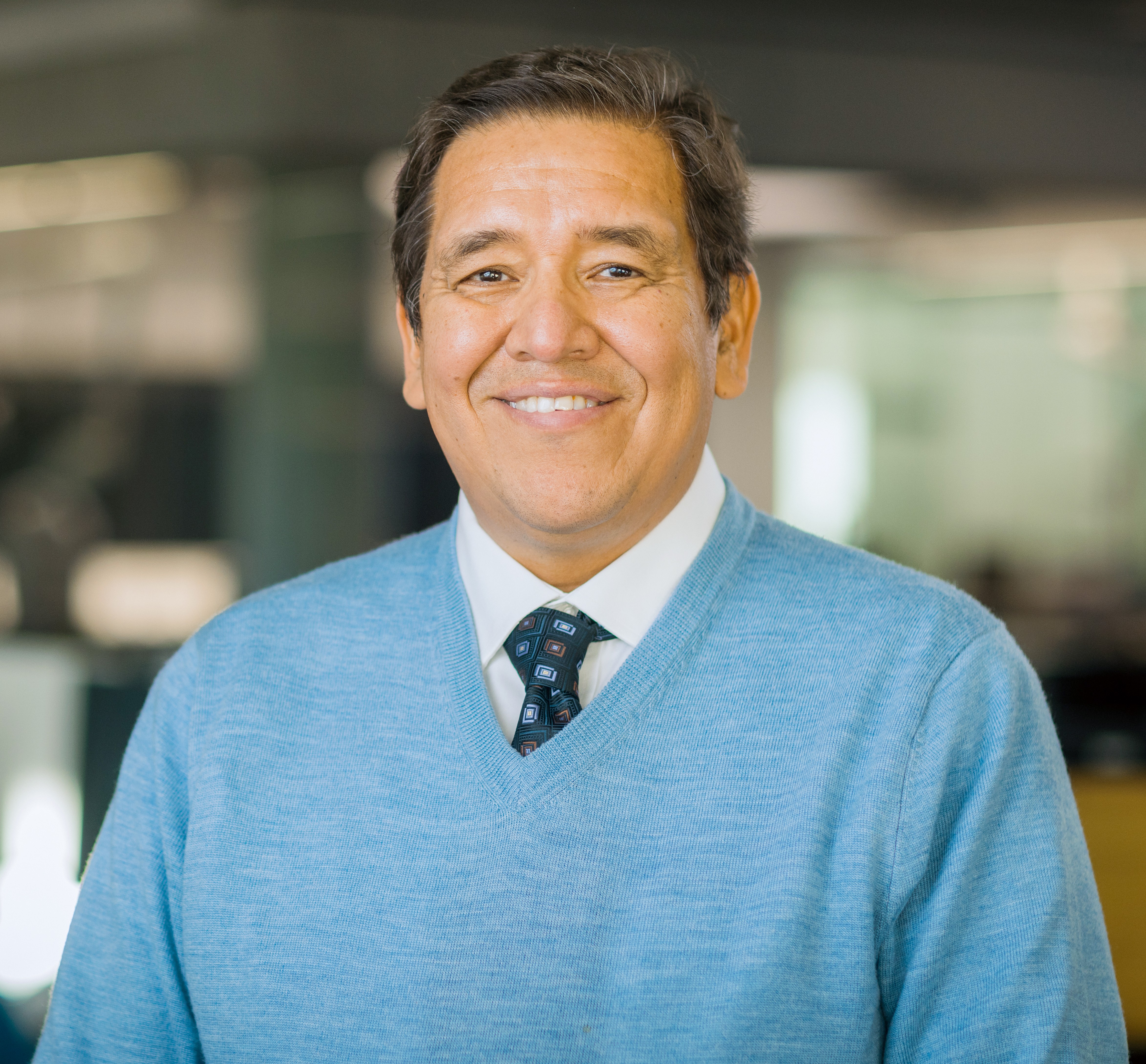 Color portrait of J.S. Onésimo Sándoval, wearing a light blue V-neck sweater over a white dress shirt and tie.