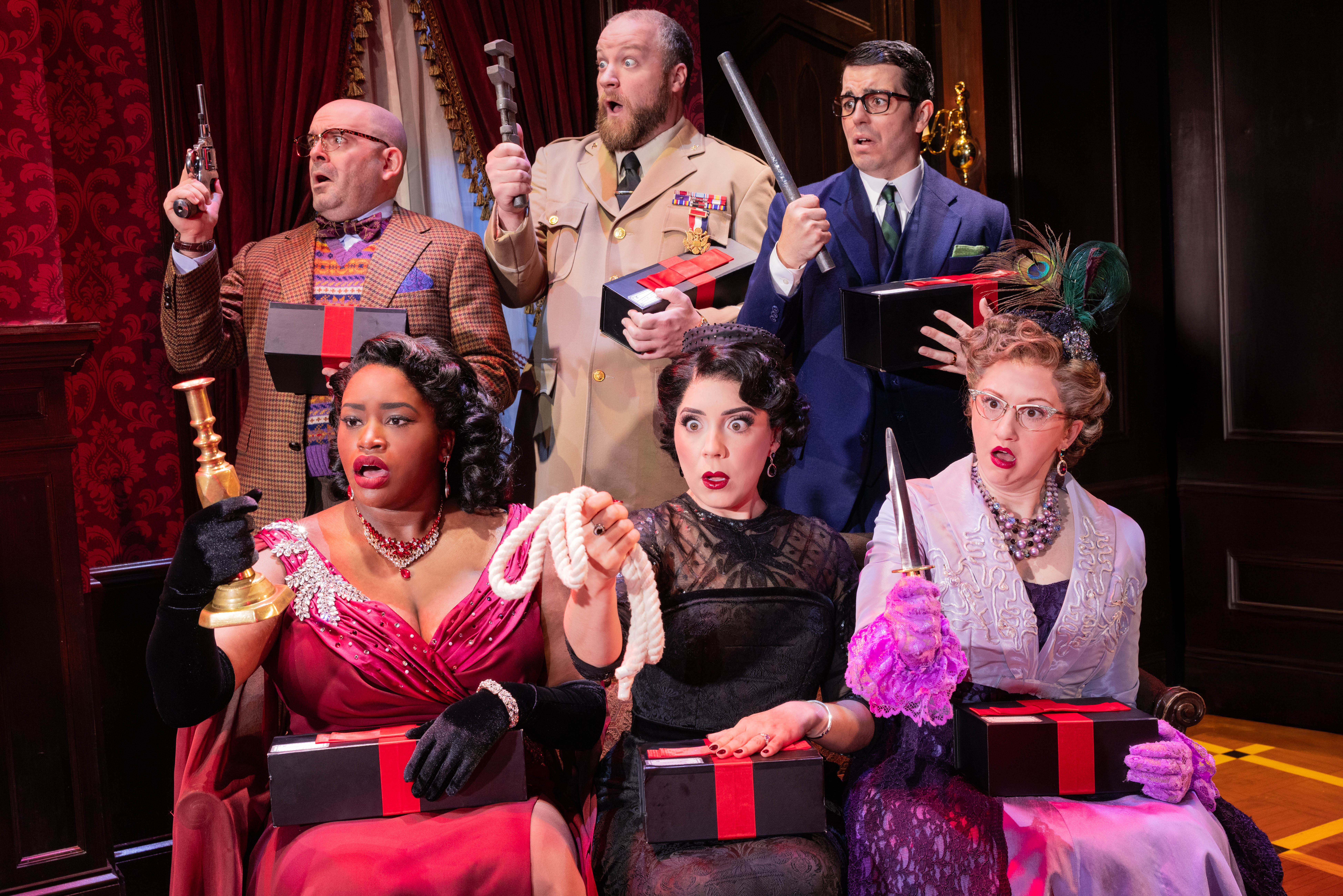 Three men and three women, all elaborately dressed, hold various murder weapons and black gift boxes with red ribbon.