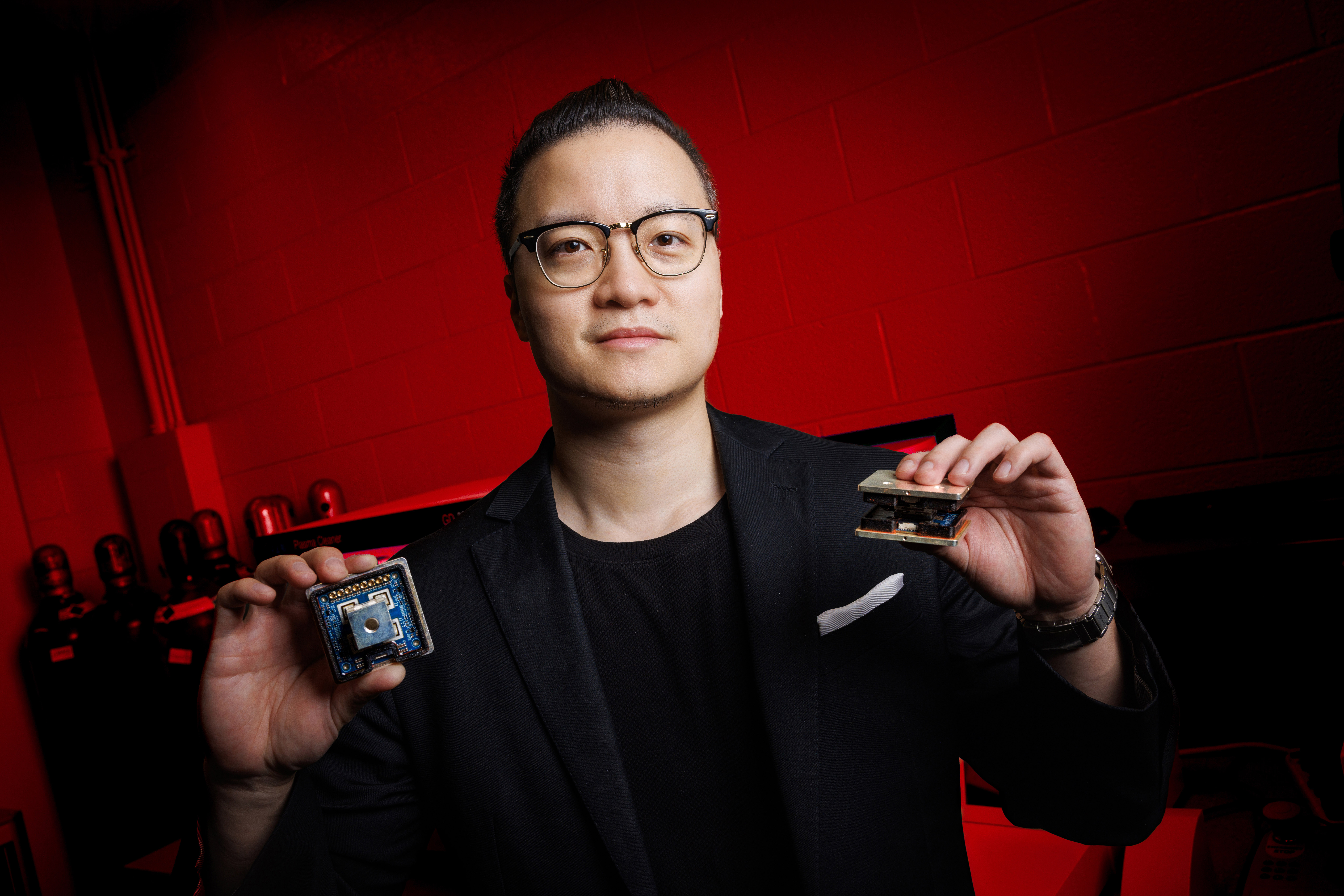 Jun Wang, assistant professor of electrical and computer engineering at the University of Nebraska–Lincoln, holds two variants of the novel 10 kilovolt silicon carbide MetaPak switch cells inside his lab in the Scott Engineering Center. He is wearing a dark blue sport coat over a black shirt and glasses. The lab is lit with red light.