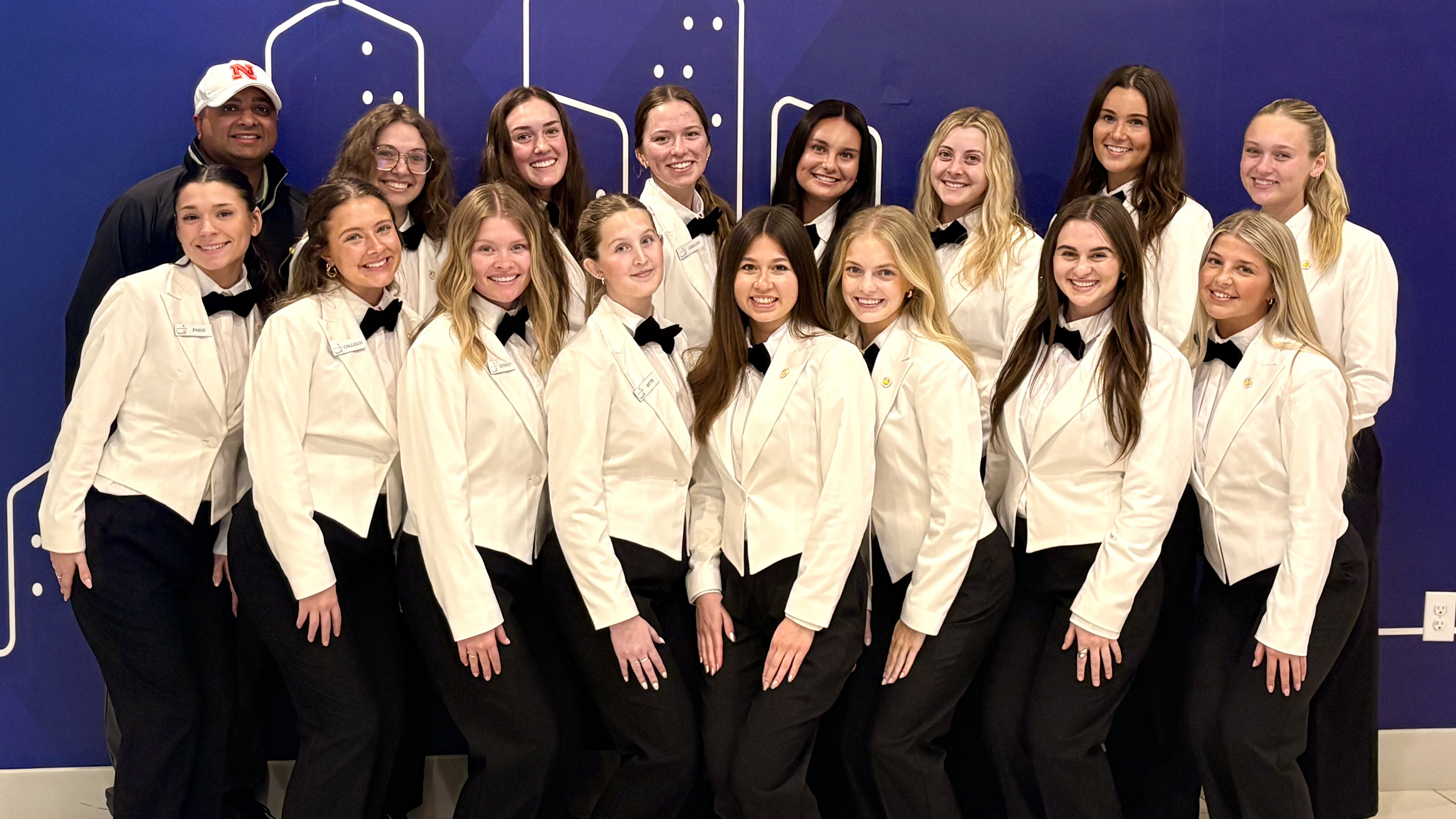 Fifteen young women wearing white jackets, white shirts, black bowties and black pants pose for a photo with instructor Ajai Ammachathram. They are Husker students providing hospitality services at the 2026 Masters Tournament.