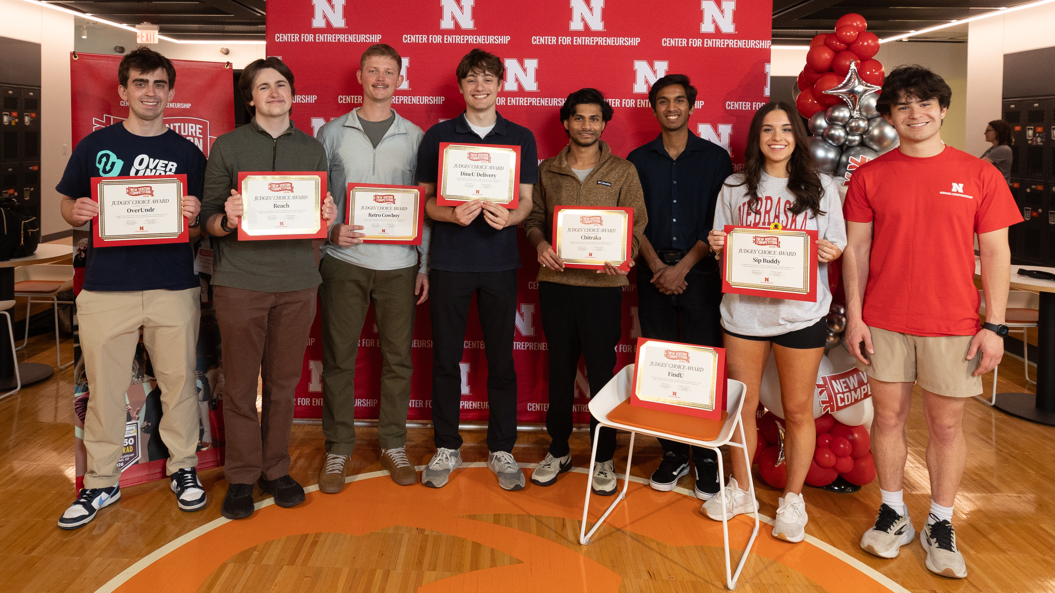 Seven young men and a young woman — all Judges' Choice Award winners in this year's New Venture Competition — hold certificates with the names of their businesses in front of a Center for Entrepreneurship backdrop and balloons.