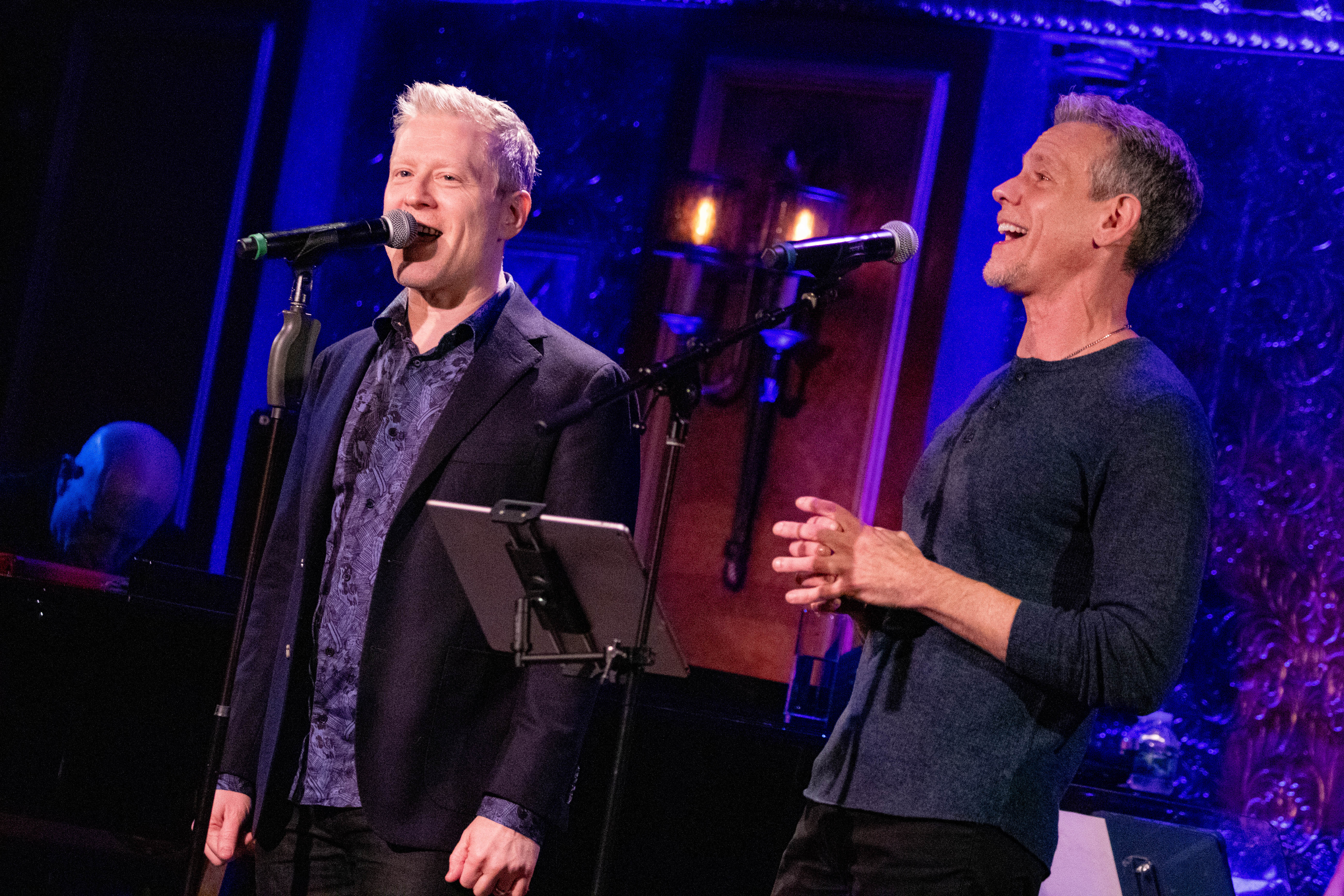 Broadway stars Anthony Rapp (left) and Adam Pascal sing into microphones on a blue-lit stage.