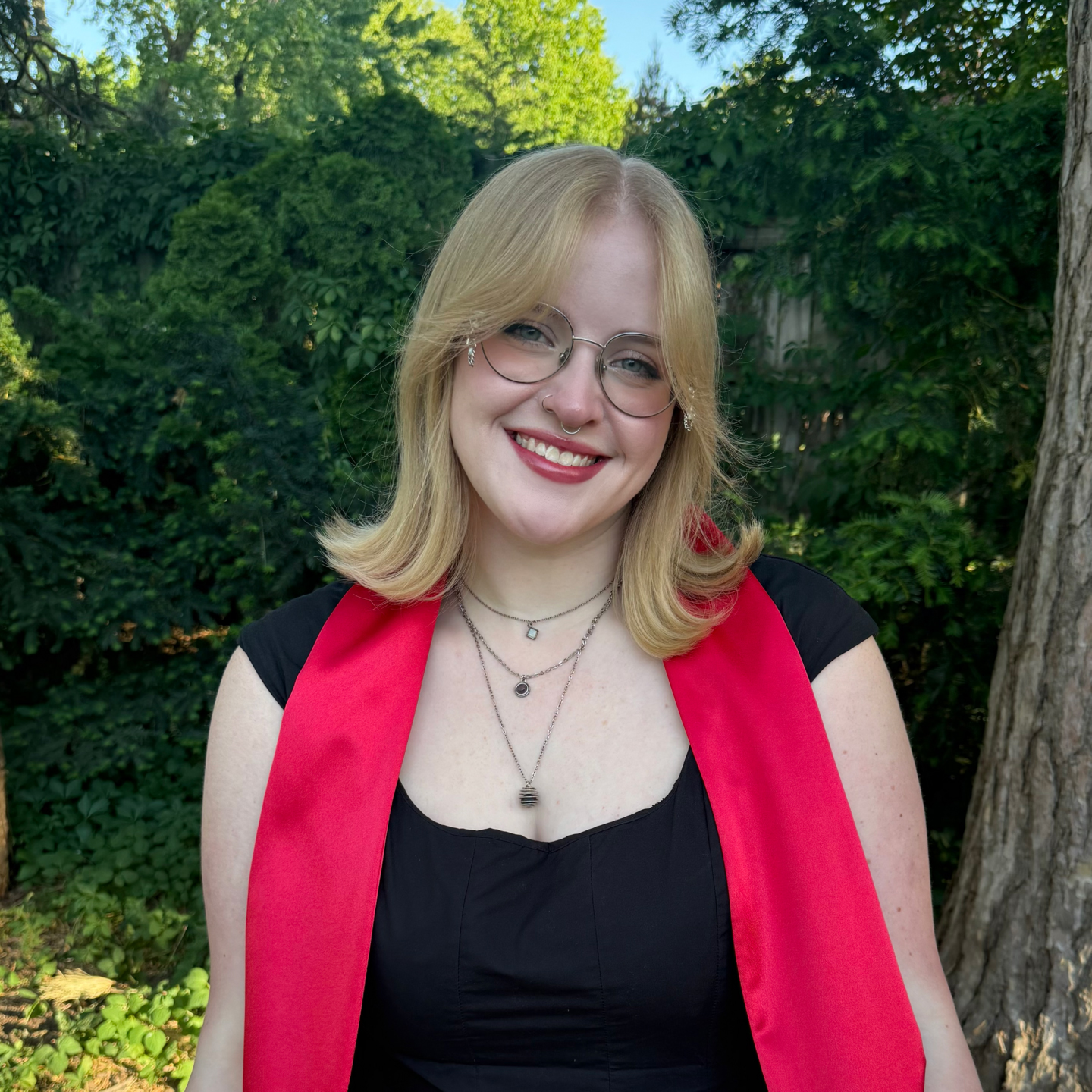 Color portrait of NSF Graduate Research Fellow Hayden Wulf, wearing a red stole over a black top with glasses and necklaces, in front of lush landscaping.