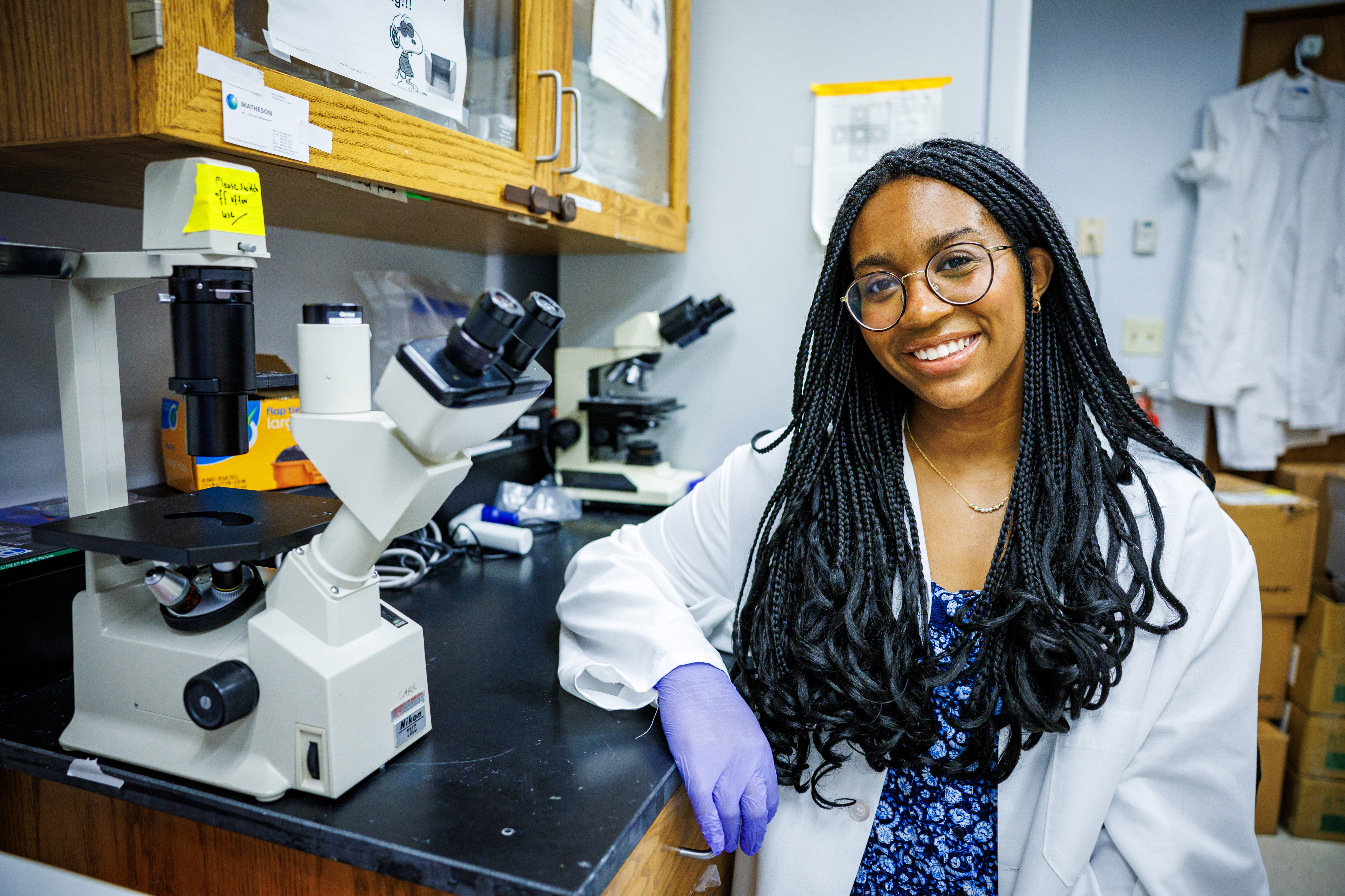 Laila Doyle, a senior nutritional science and dietetics major at the University of Nebraska–Lincoln, poses next to two microscopes in a lab. She is wearing a white labcoat over a blue floral dress, with purple protective gloves and glasses.