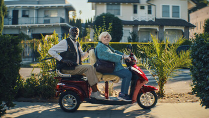 Scene from the film "Thelma" with two elderly people sitting on a motorized cart on a sidewalk in front of homes.