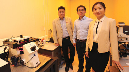 UNL engineers (from left) Yongfeng Lu, Lijia Jiang and Ying Liu stand in a laboratory illuminated by a special light designed to protect the microscope at left.