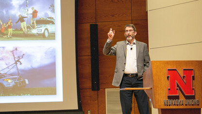 Adam Houston speaks behind a lectern with a screen showing storm-chasing photos behind him.