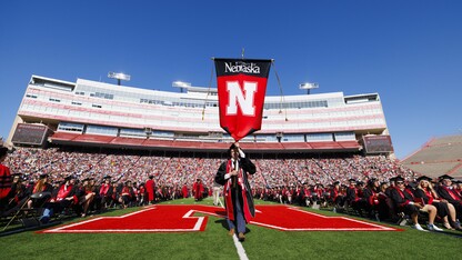 Graduate Paul Pechous carries a banner past rows of seated graduates on the field at Memorial Stadium.