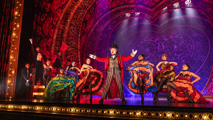 A man in circus attire opens his arms wide in front of eight women in colorful burlesque outfits and three men in tuxedos on stage.