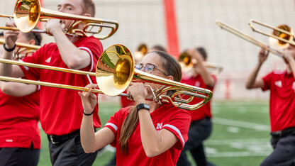 Young men and women in red shirts and black pants play trombones on a football field.