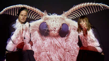 The Moth Project's Peter Kiesewalter and Whitney La Grange, dressed in white suits, sit next to a large projected image of a moth.