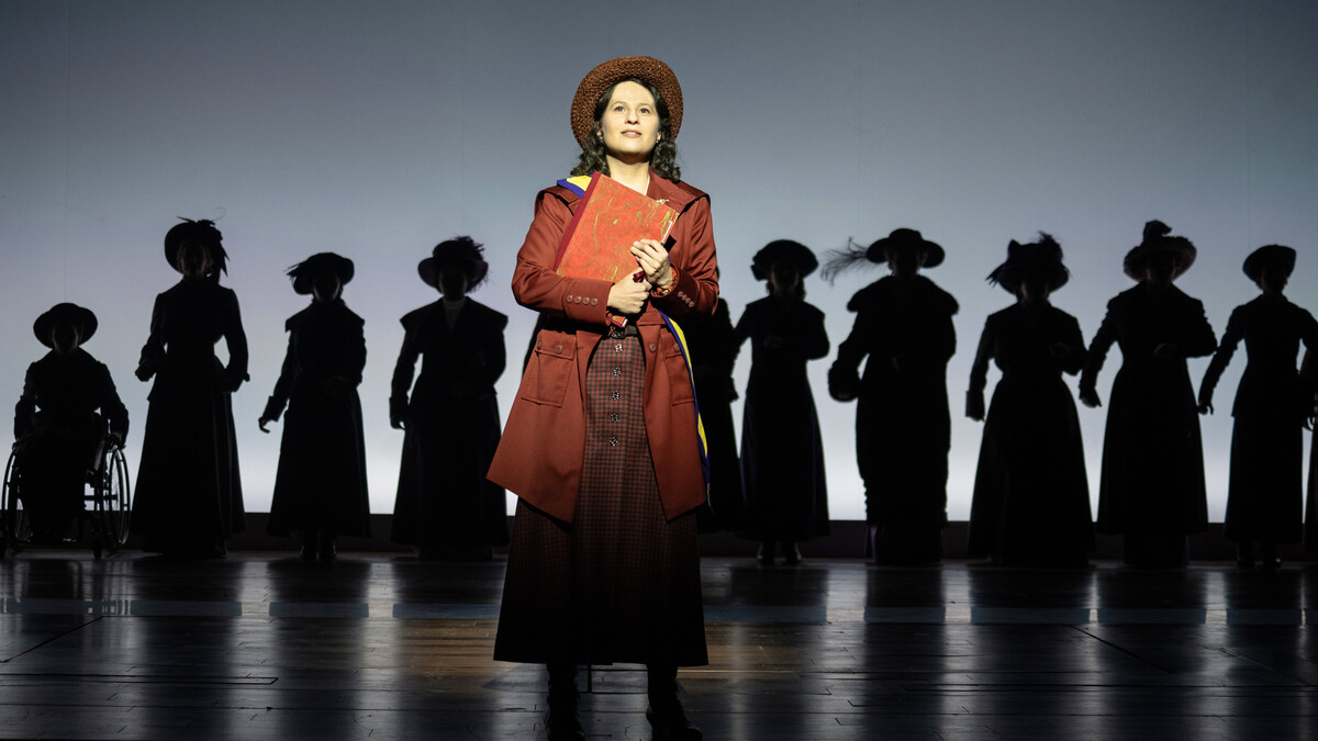 Shaina Taub (foreground) stars as women's rights activist Alice Paul in the musical "Suffs." She is seen in a maroon dress, overcoat and hat, holding a book, in front of a line of other suffragettes in silhouette.