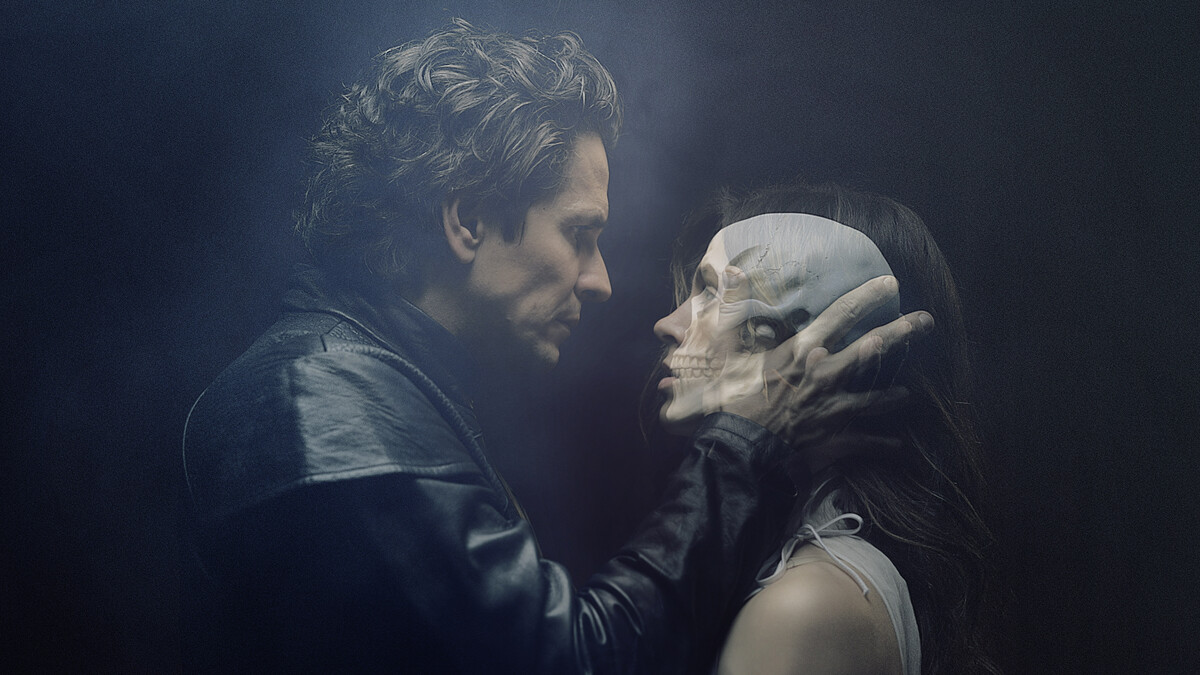 A man and woman stare at each other as he holds her head in his hands. A skull image shines through the woman's head.