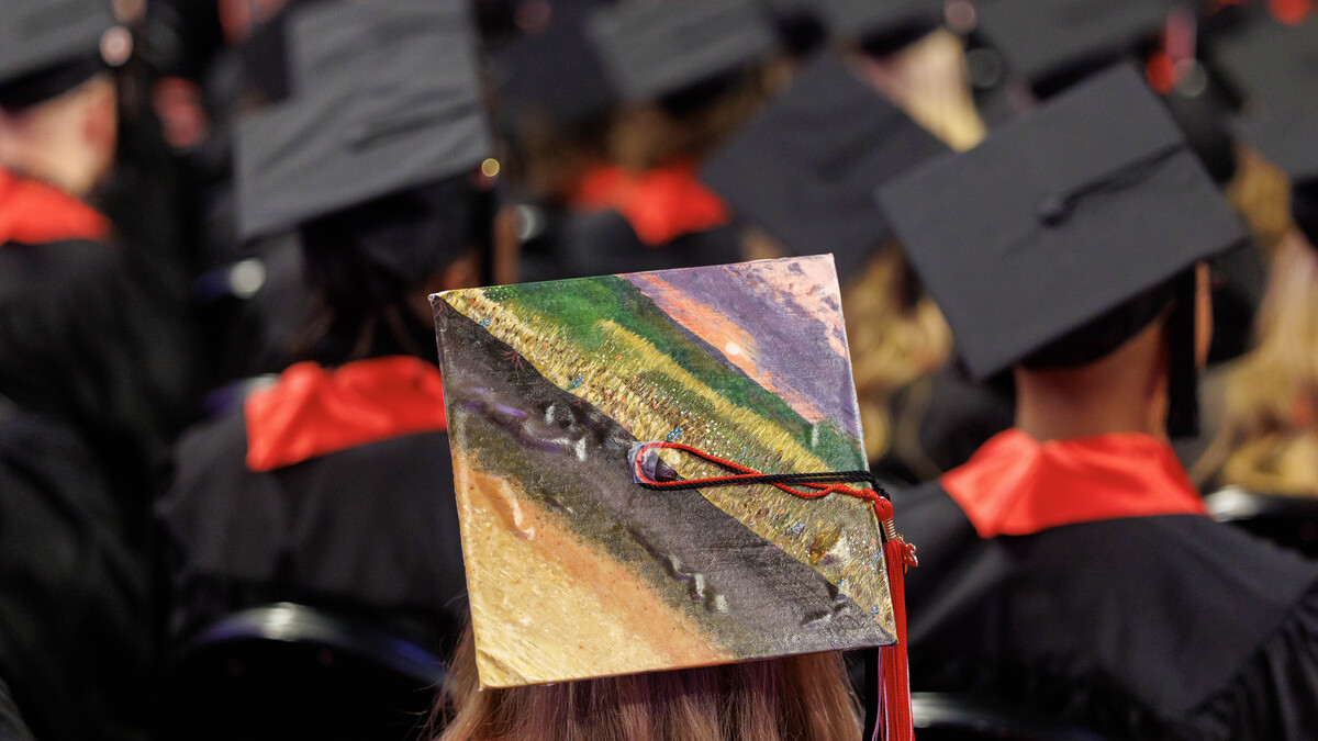 A mortar board is decorated with a field at sunset among seated graduates.