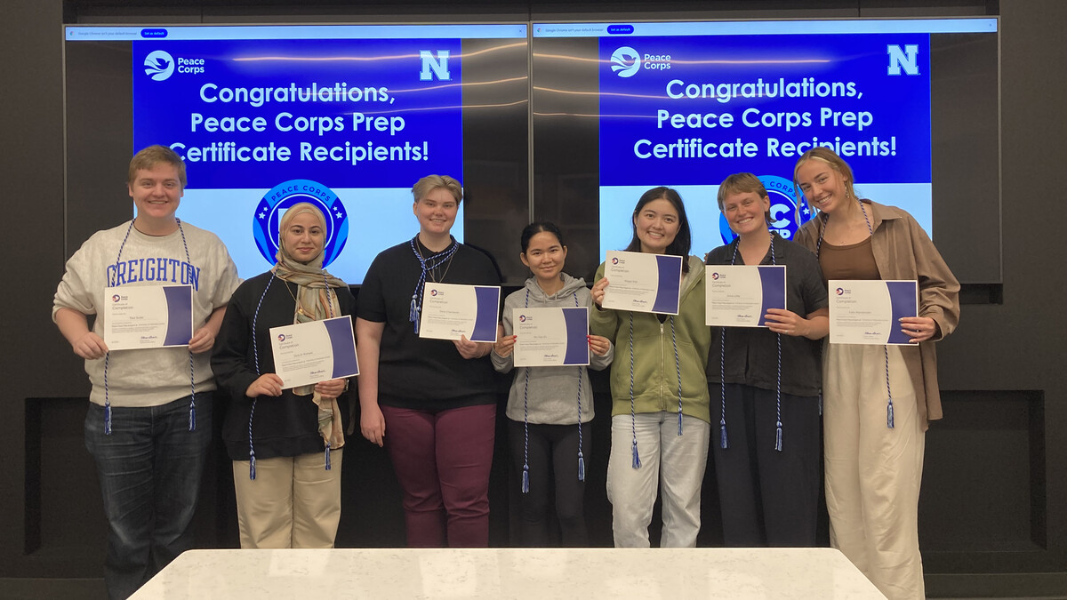 Seven University of Nebraska–Lincoln students stand in a row, holding certificates. Behind them are two screens that say "Congratulations, Peace Corps Prep Certificate Recipients!"