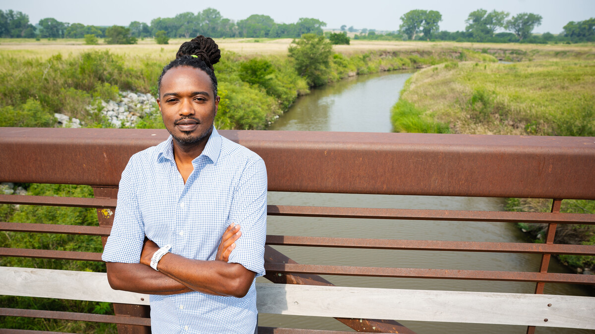 Ng’ang’a Wahu-Mũchiri, associate professor of English, stands on a bridge, over a river, with his arms crossed. His is wearing a blue-and-white plaid shirt.