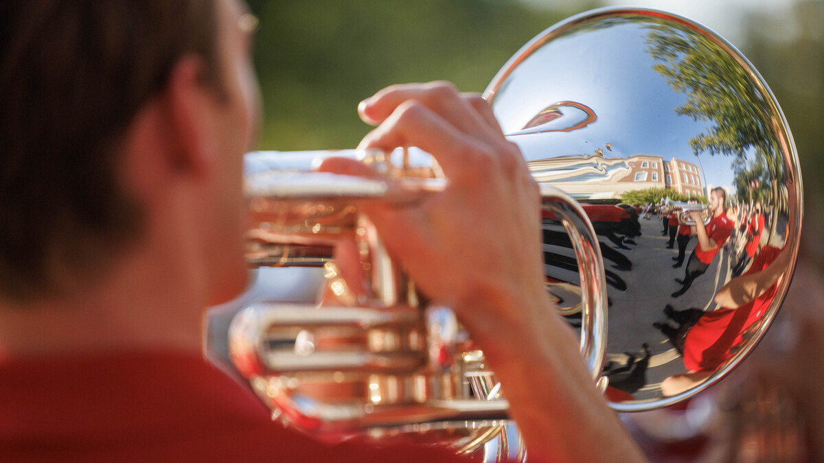A brunette man plays a french horn, where other marching band members can be seen in the distorted reflection of the brass horn.