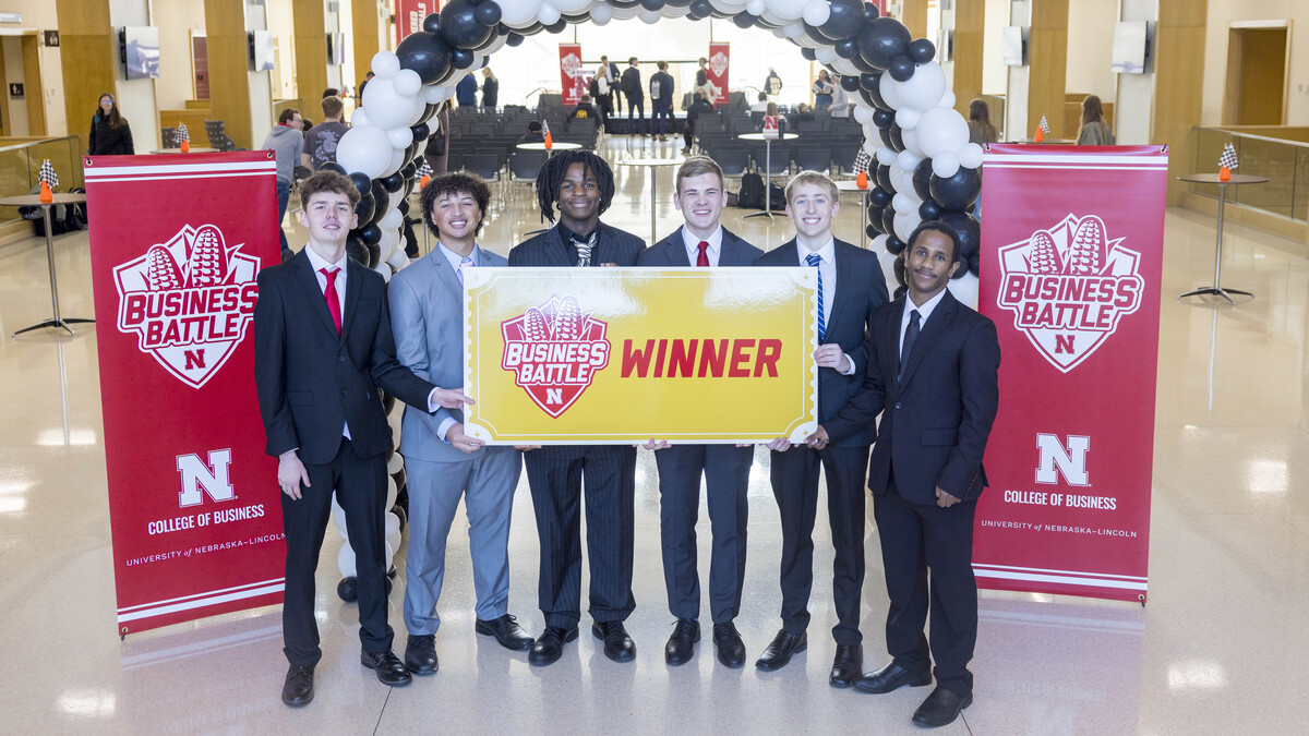Six young men in suits pose with a poster that reads "Business Battle Winner" in the Howard L. Hawks Hall atrium.
