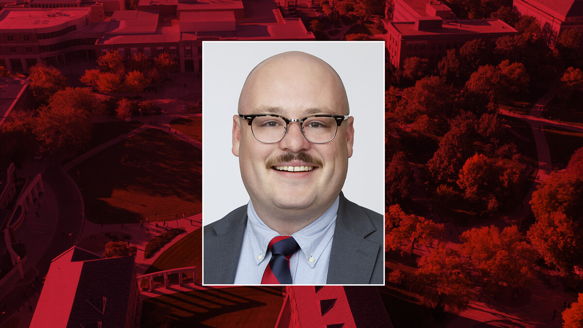 Color portrait of Julian Gillilan, doctoral candidate in the University of Nebraska–Lincoln's Department of History, on a red campus background. He is wearing a dark gray sport coat over a light blue dress shirt and red-and-blue striped tie, with glasses.