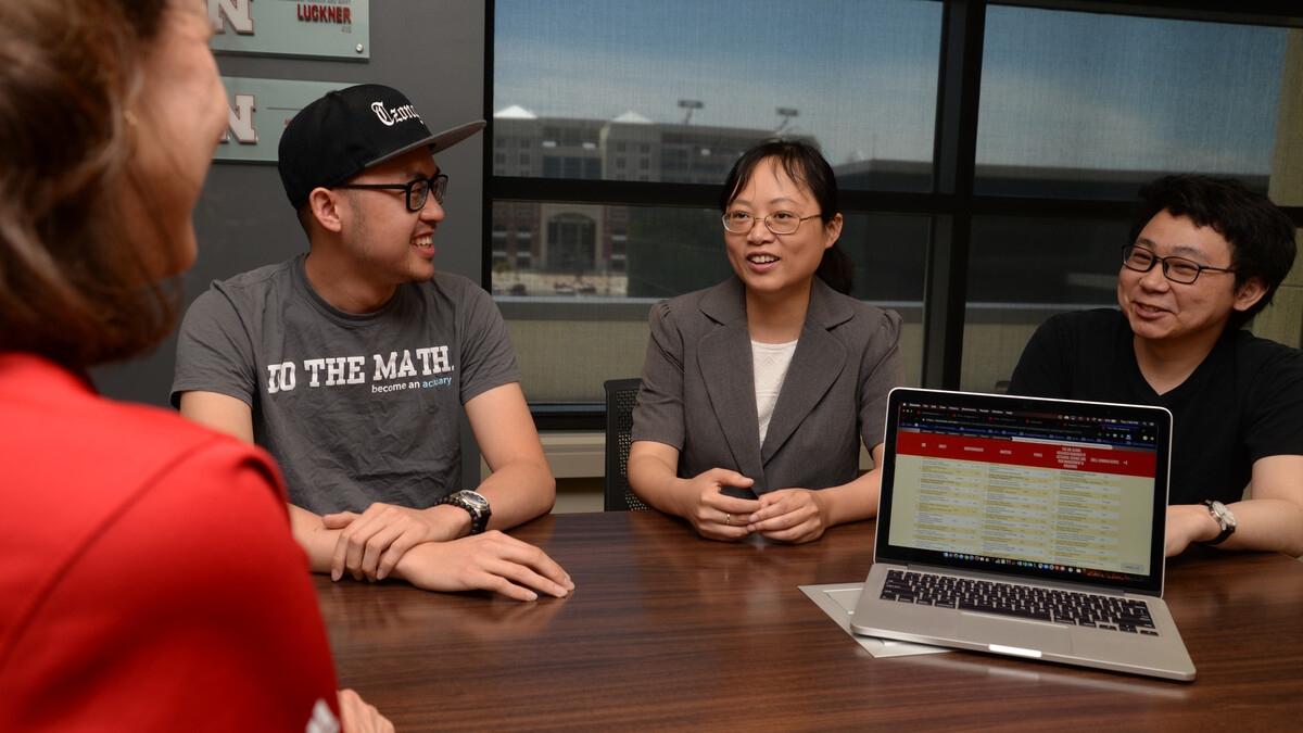Two men and two women sit around a table, with an open laptop on it. Memorial Stadium can be seen through a window in the background.