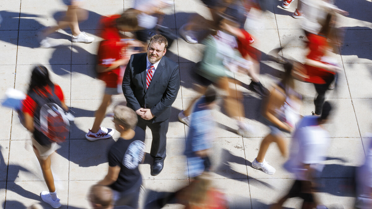 A man in a black suit with a red tie stands in the middle of a blurry people walking around him.