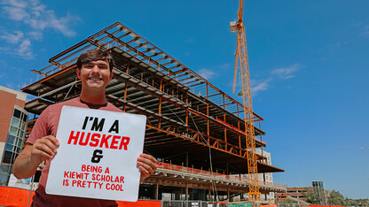 Kiewit Scholar Maverick Naughtin stands outside the Kiewit Hall construction site holding a sign that says, "I'm a Husker and being a Kiewit Scholar is pretty cool."
