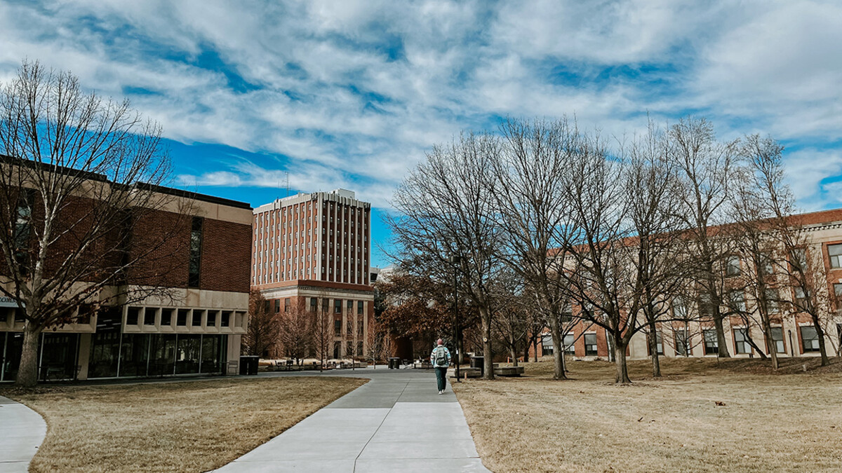 A student walks down a concrete path with three red brick buildings in the background.