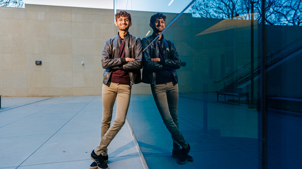 Aaryan Naik leans against Hawks Hall for a photo with his reflection in the glass of the building