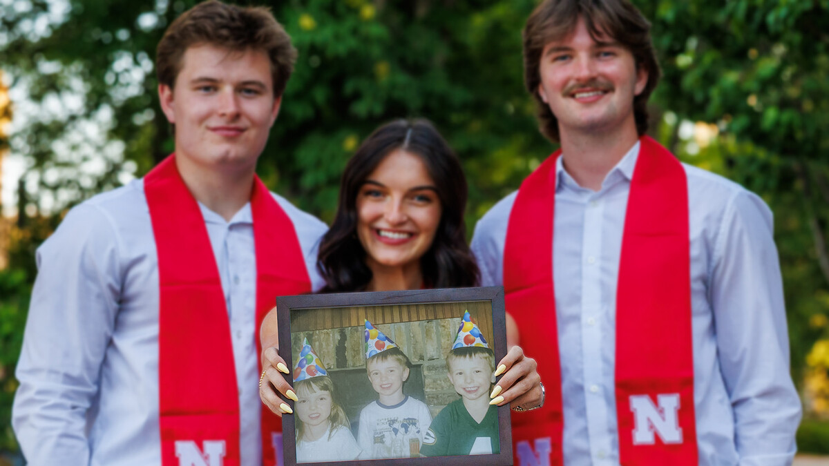 Two men in white button-ups smile next to a brunette woman holding a childhood photo of all of them in party hats.
