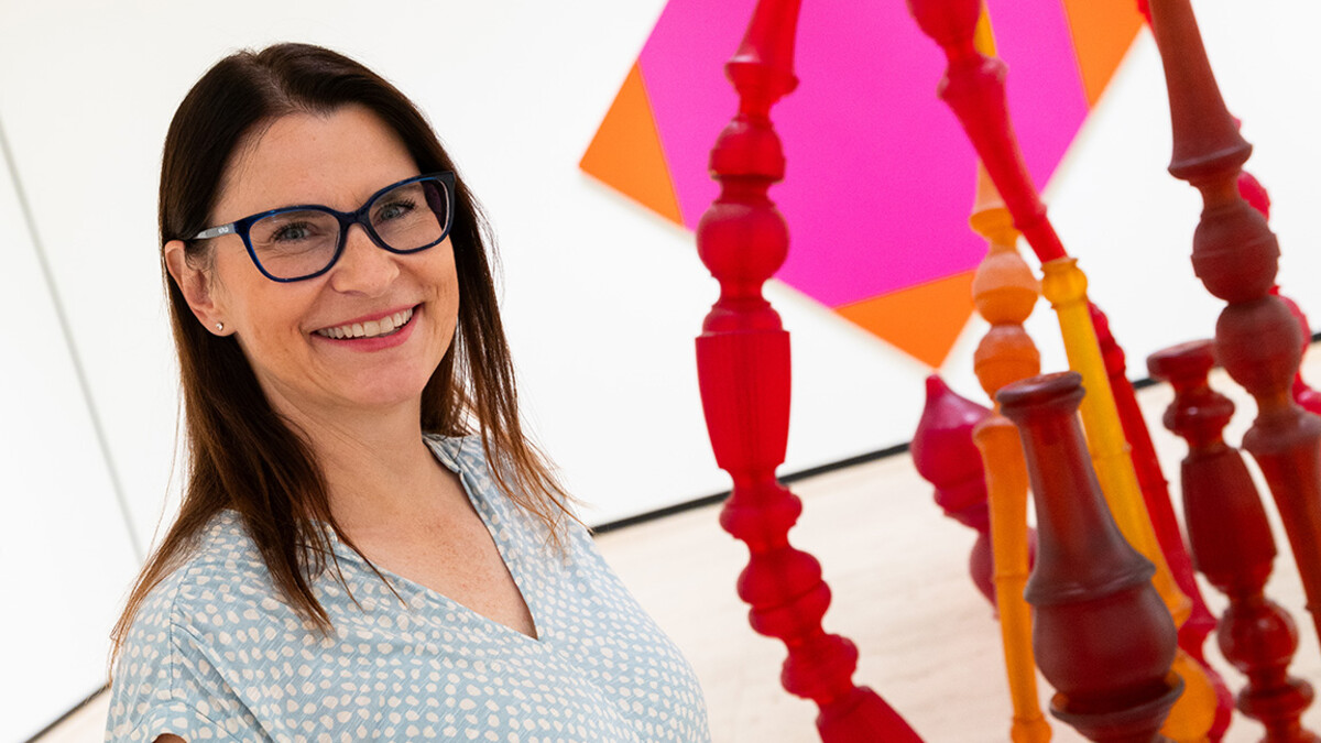 A white, brunette woman with black glasses and a light blue blourse stands next to a sculpture of multiple red, yellow and orange oddly-shaped tubes.