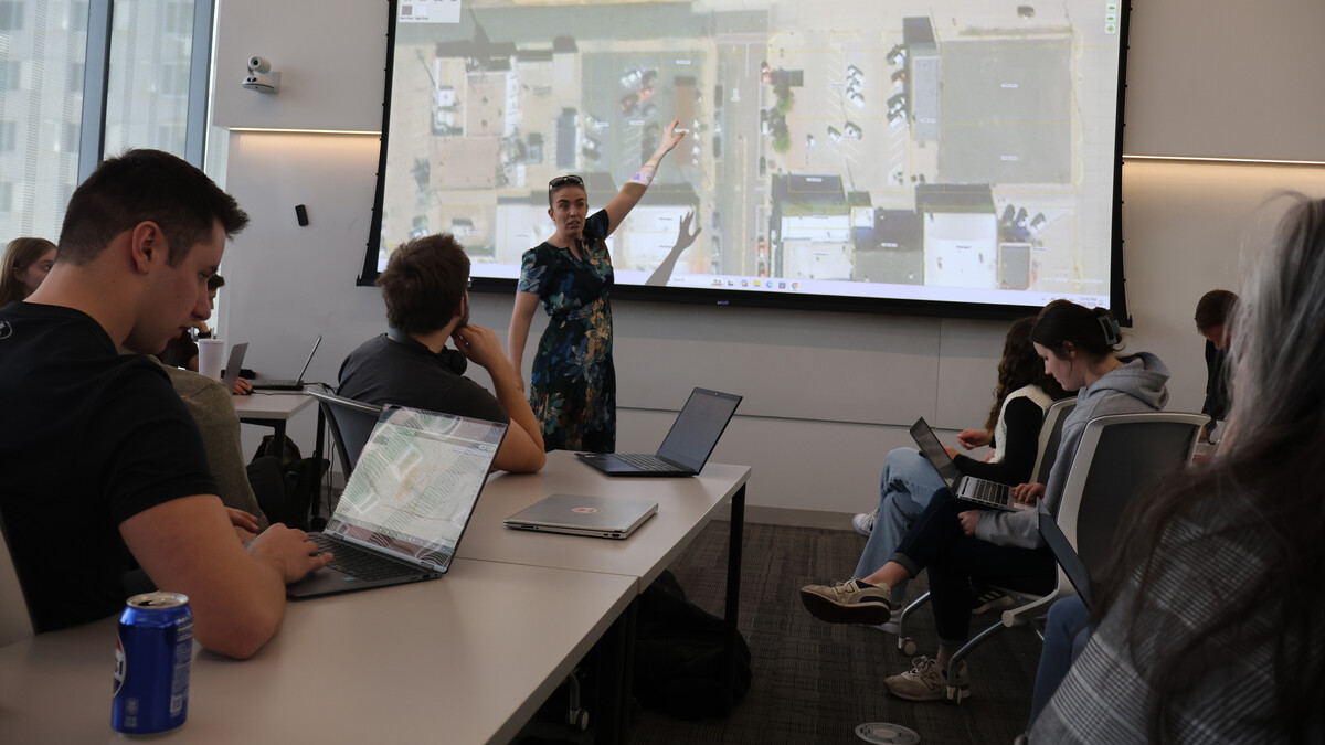 Students gather in front of a projector as a woman in a floral dress goes through maps of an aerial view of downtown Sydney, Nebraska.
