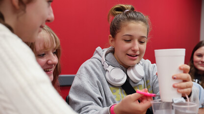 Lyla Mayle sits with her brown hair in a bun wearing a grey sweatshirt with white headphones around her neck, cutting a foam cup with a pair of scissors.