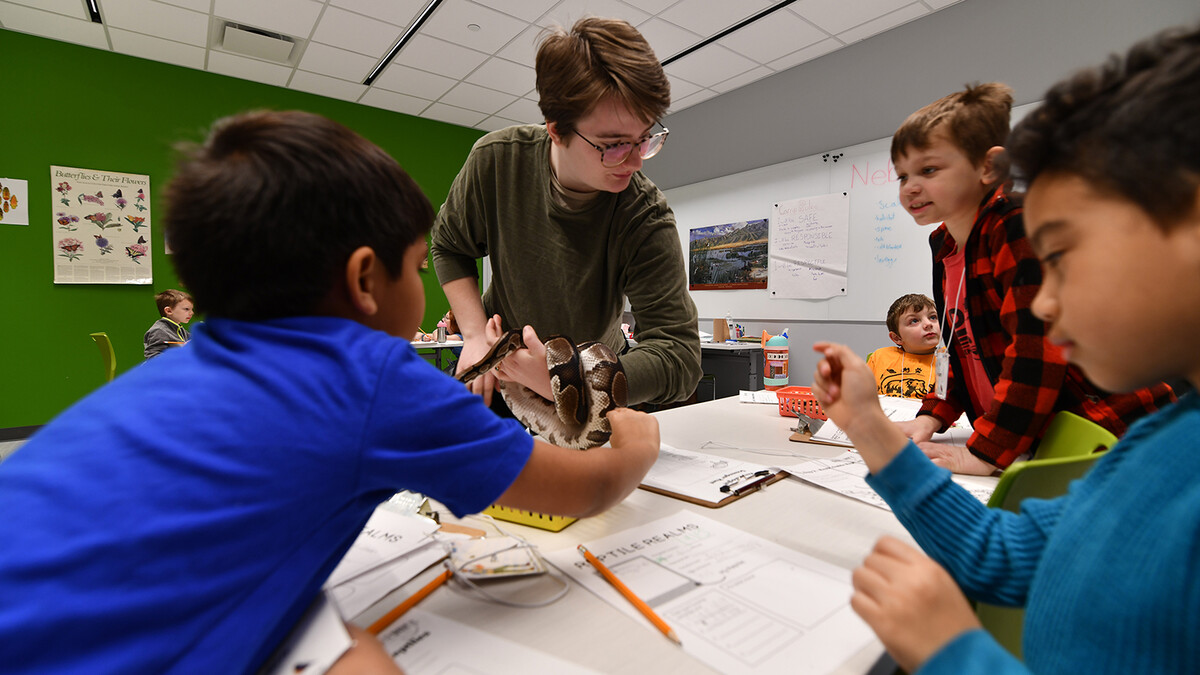 kids gather around a table of textbooks with an adult man, short brunette hair with glasses, in a green sweater.