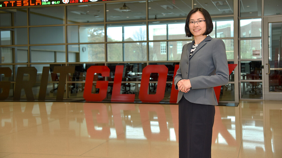 A brunette woman with a bob stands with her hands together, wearing a grey blazer, black slacks and thin-framed glasses.