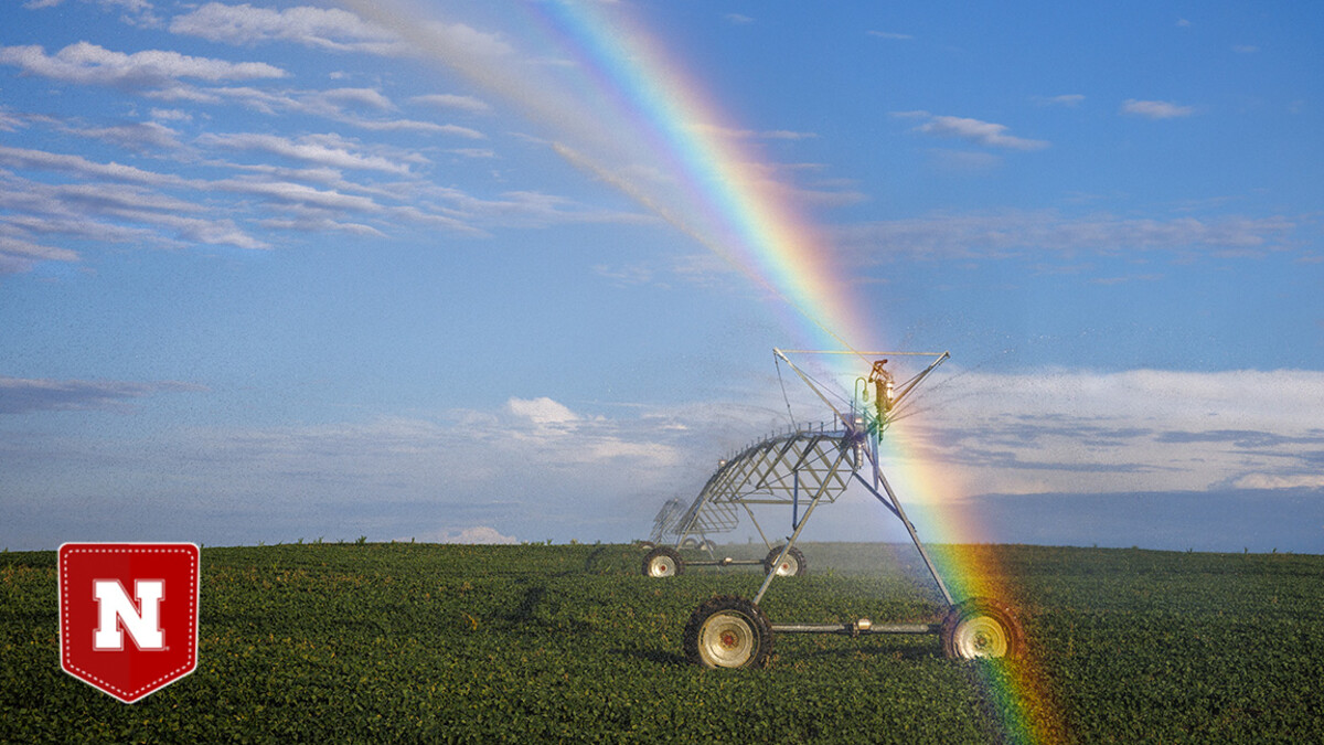A rainbow forms over an irrigation pivot in a grain field, with a blue sky in the background.