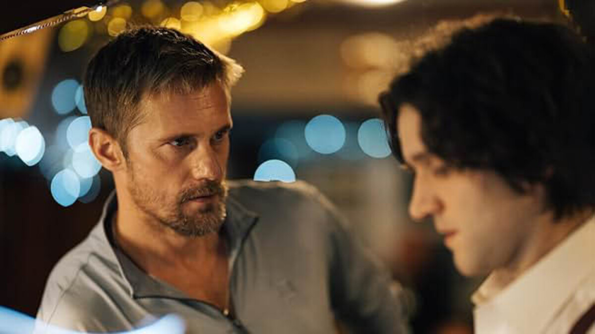A still image of "Pillion," where a blond man with short hair and a beard stands in a grey sweater next to a brunette man with hair reaching his shoulders in a white button-up.